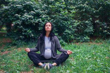 Pregnant young woman practices yoga in summer park, copy space. Preparation for childbirth, breathing technique