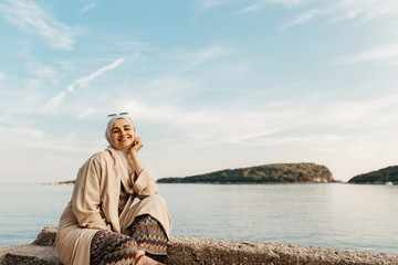 Fototapeta premium portrait of young European Muslim women with hijab sitting on the stone beach with sea in the background. She is happy and relaxed.