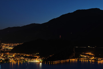 Fototapeta na wymiar Evening panoramic view of the of Riva Del Garda, Lake Garda. City night lights reflected in water. Aerial view of the city of Riva del Garda