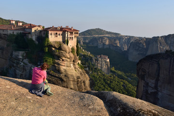 Sitting on the rock woman takes photo of Varlaam Monastery at sunset, Rosanou nunnery seen at distance. Meteora - UNESCO world heritage site, Greece 