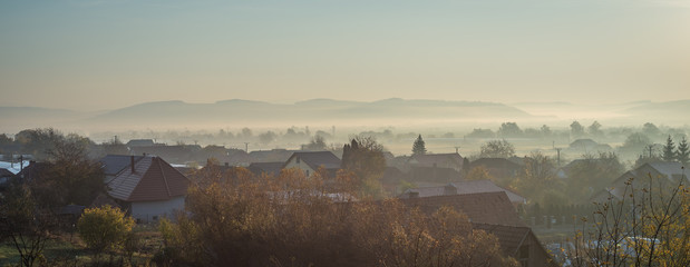 Panorama of morning fog in a valley. Scenic silhouettes of trees and buildings.