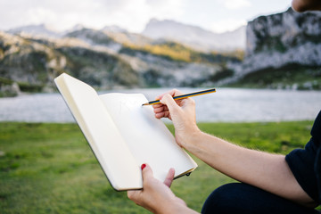 A beautiful caucasian blonde woman writes and draws on a notepad in a mountain landscape with lake