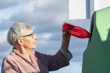 older woman putting a red glass bottle in a Green glass recycling container