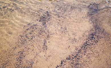 Background of golden river sand mixed with small black pebbles, a small water ripple is visible on the surface of the water; texture