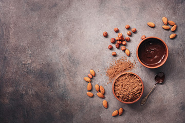 A bowl of melted chocolate, cocoa powder, almonds and hazelnuts on a dark rustic background. Top...