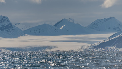 Snow covered mountains and a glacier by an arctic fjord with waves gleaming in the sun