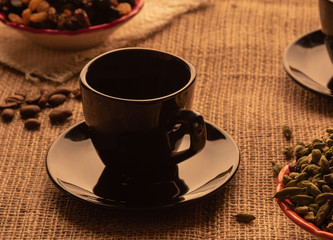 black coffee cup and cardamom grains on a table with burlap on the background of coffee beans and raisins
