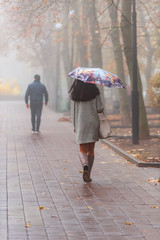 Morning fog in town in the fall. A woman with a colored umbrella is walking along the alley. Concept - urban clothing style.