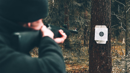 A man with a black air rifle in his hands shoots at a target fixed on a tree trunk in the forest,...
