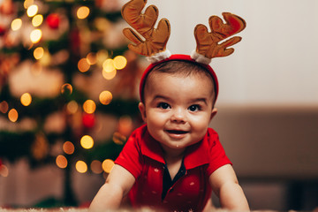 Cute baby boy wearing reindeer antlers crawling on floor over Christmas lights. Looking at camera....