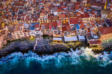Obraz premium Aerial view of the rocky coast and the houses of Cefalu from the sea in windy and stormy weather. Big waves beat against stones. Streets with the city lights. Italy, Tyrrhenian SeaSicily, Italy