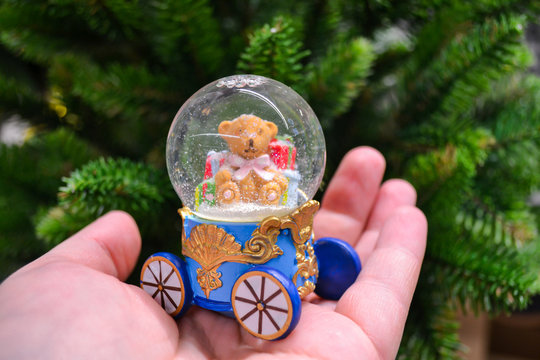 The hand holds a snow globe in the form of a carriage, inside which sits a Teddy bear with gifts, and snowflakes are spinning. On the background of green fir branches