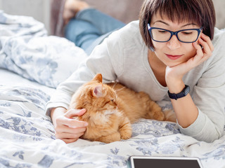 Cute ginger cat and woman in glasses are lying in bed. Woman is reading tablet and stroking her fluffy pet. Morning bedtime.