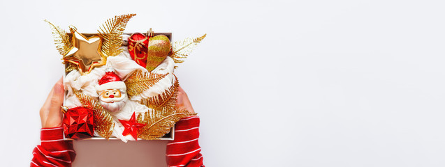 Woman in red sweater is holding white box with New Year decorations. Bright red and golden decorative toys for Christmas tree. Top view, flat lay.