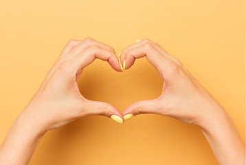 Fototapeta premium Female hands show a heart symbol isolated. Bright manicure. Warm yellow background