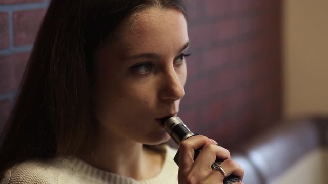 Vaping teenager. Young pretty white caucasian teenage girl with problem skin smoking an electronic cigarette indoors. Life-threatening bad habit. Vape activity.