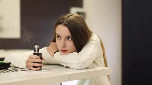Vaping teenager. Young pretty white caucasian teenage girl with problem skin smoking an electronic cigarette indoors. Life-threatening bad habit. Vape activity.