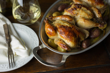 Close up view of roasted Cornish hens with small fingerling potatoes, shown with vintage fork, small white plate, stemless glass of white wine and cloth napkin. 