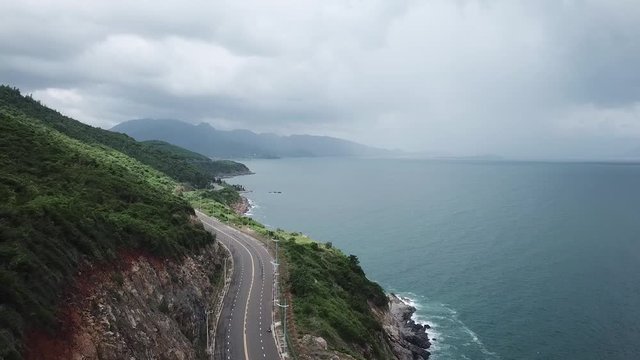 A flying camera takes a motorcyclist passing on a road near the sea with a view of the rocky shore.
