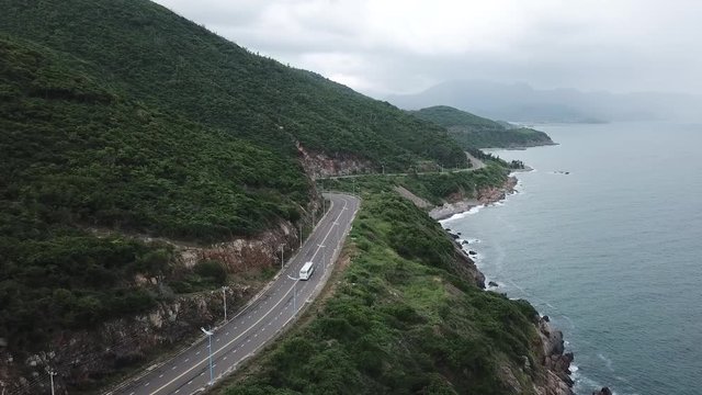 A flying camera takes a motorcyclist passing on a road near the sea with a view of the rocky shore.