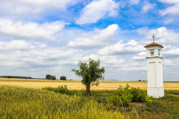 Fototapeta na wymiar A little Christian Chapel in the middle of Upper Austria rural field with golden yellow grain during a beautiful sunny day under blue sky with white fluffy clouds
