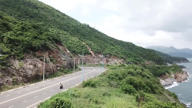 A flying camera takes a motorcyclist passing on a road near the sea with a view of the rocky shore.