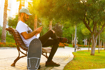 young man sitting on bench looking at cellphone with skate with sunset light