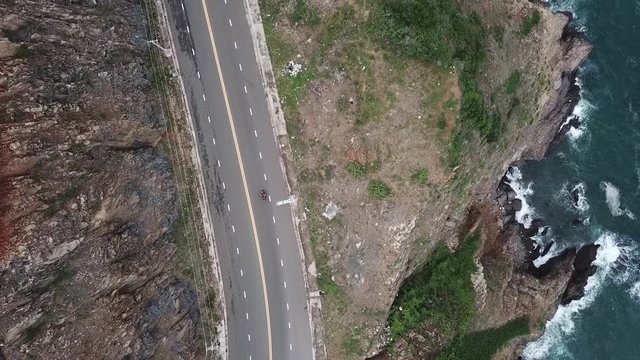 A flying camera takes a motorcyclist passing on a road near the sea with a view of the rocky shore.