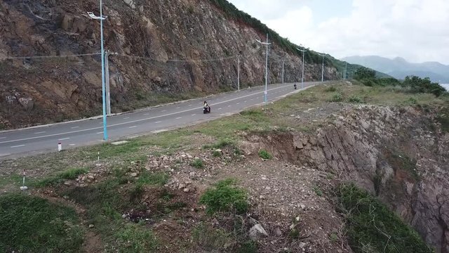 A flying camera takes a motorcyclist passing on a road near the sea with a view of the rocky shore.