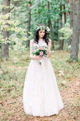 Fototapeta na wymiar Beautiful bride with eucalypthus wreath on the head standing in the forest and looking at wedding bouquet of different flowers. Rustic style