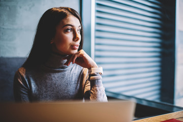 Dreaming attractive female freelancer looking in window and thinking about weekend in break of work on modern laptop computer indoors,thoughtful hipster girl sitting desktop with new advanced netbook
