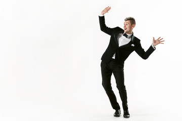Portrait of young smiling handsome show man in tuxedo stylish black suit, studio shot dancing at white background. Businessman in jacket with bowtie