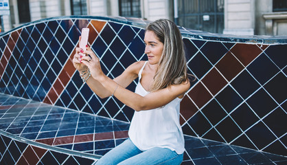 Attractive Caucasian hipster tourist enjoying Spanish holidays during summer sitting on ceramic bench and photographing landscape of city, beautiful woman in casual wear clicking pictures for memorise
