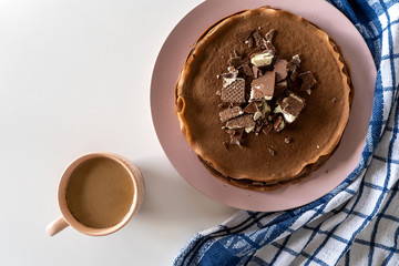 Сhocolate pancakes with chocolate and waffles on a pink plate, a cup of coffee and blue towel on a white table. Traditional breakfast. Top view