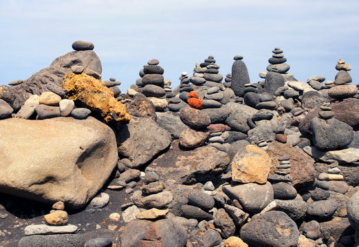 close up of towers of stacked pebbles and colored stones in a large arrangement on a black sand beach with blue sky