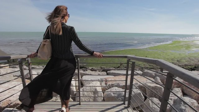 Woman with black dress moved by the wind watching the sea on the horizon. Concept: wanderlust, travel lifestyle. Caorle, Venice, Italy