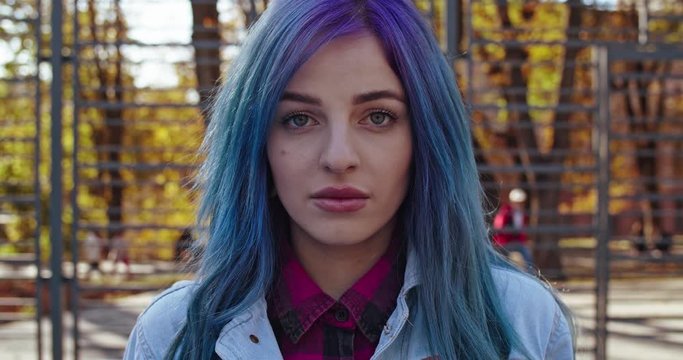 Portrait of the young Caucasian hipster girl with blue hair looking straight to the camera in the park in fall. Outdoor. Close up.