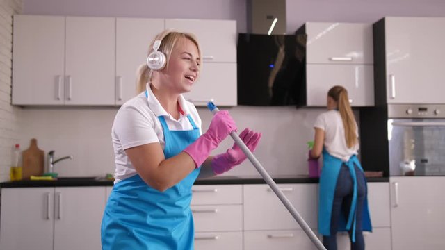 Close-up of happy female cleaner in headphones singing and dancing while washing floor in home kitchen, her workmate wiping surfaces on background. Professional janitors during house cleaning