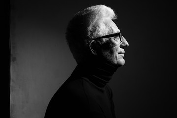 Never get old, handsome and sexy at every age concept. Close up portrait of fashionable mature man wearing trendy eyewear, black turtleneck, sitting in art gallery. Modern haircut. Silver hair