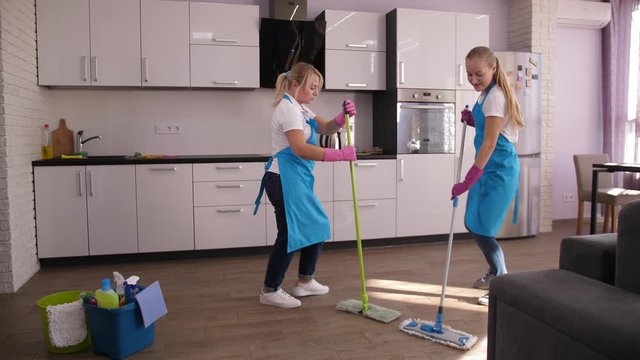 Cheerful pretty cleaners in aprons and gloves dancing with mops while washing floor in house kitchen. Happy adult women, workers of cleaning service enjoying fun and dance during domestic wet cleaning