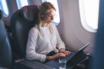 Caucasian female working on freelance during flight and using comfortable service of wireless internet access on board. Young blonde hair woman listen to music, keyboarding email on tablet in airplane