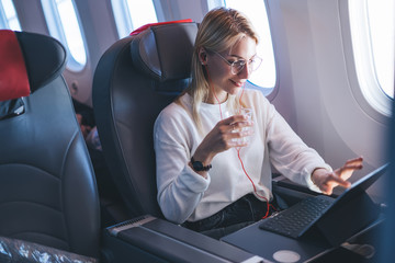 Smiling attractive female passenger watching in earphones online movie on touch pad and during...
