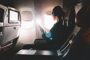Female tourist in headphones listening audio book and looking in airplane window enjoying flight. Young 25 years old woman with cellular phone in hands sitting on seat during flight in first class