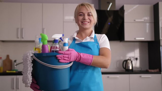 Positive female cleaner in apron and gloves posing with bucket full of detergents in kitchen shining with cleanliness. Blonde worker of cleaning company smiling while looking at camera after clean up