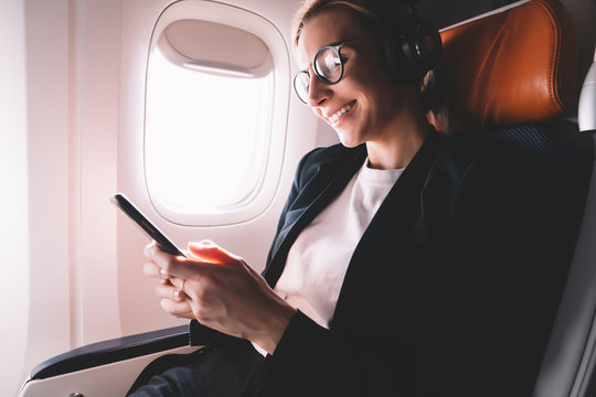 Cheerful female passenger in eye wear feeling happy from received message with good news connected to wireless internet on board, smiling young woman in headphones for noise cancellation