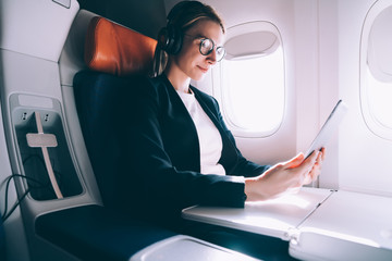 Caucasian woman in eyeglasses reading news on financial website during intercontinental flight in airplane while listening audio book in earphones for noise cancellation, female watching video
