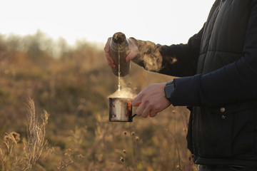 A woman pours a hot drink from a thermos into a mug. Refreshment while hiking. Clink glasses of tea in the morning at dawn, three people drink tea. Metal cups on the nature. Hands warm hot coffee