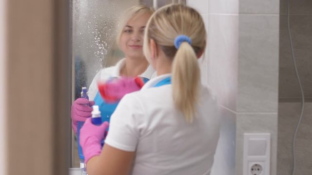 Back view of pretty maid spraying detergent and wiping mirror in hotel bathroom during clean up. Blonde cleaning lady smiling while looking at her reflection in clean shiny surface of bathroom mirror