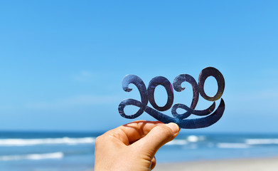 Happy New Year creative concept, hand holding 2020 text against blue sky and beach blur defocused background.