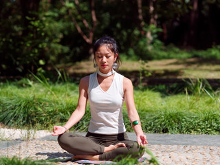 Beautiful Chinese woman wearing sport clothes practices yoga and meditates in the lotus position on mat with defocused background, young female doing yoga asana in the park with eyes closed.